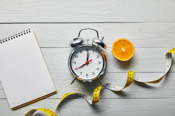 Top view of orange half, blank notebook and measuring tape near alarm clock on wooden white background — Stock Photo