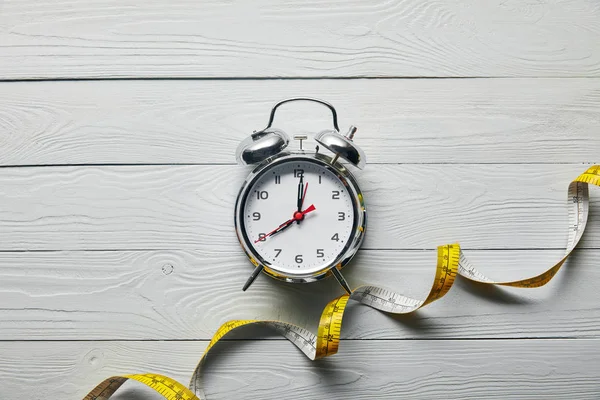 Vista superior del reloj despertador y cinta métrica sobre fondo blanco de madera - foto de stock