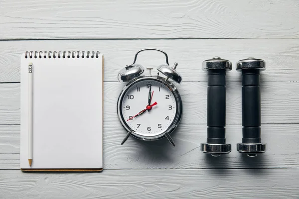 Tendido plano con despertador de plata, cuaderno vacío con lápiz y mancuernas sobre fondo blanco de madera - foto de stock