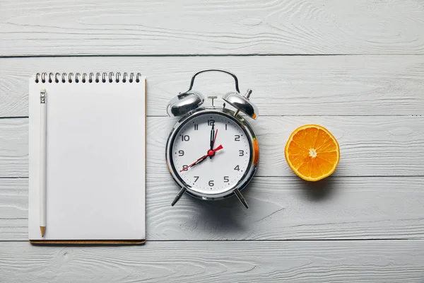 Tendido plano con despertador de plata, cuaderno vacío con lápiz y medio naranja sobre fondo blanco de madera - foto de stock