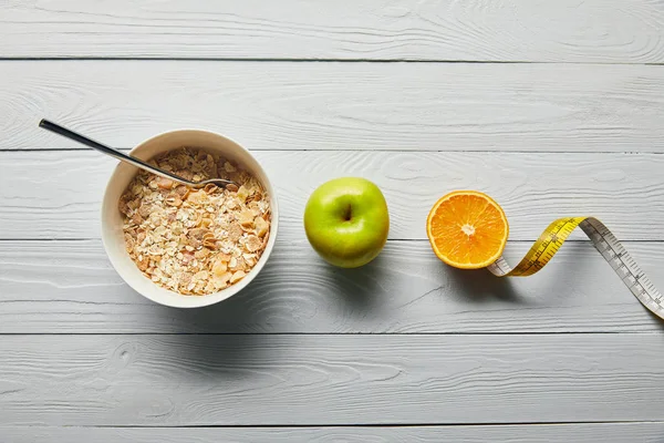 Flat lay with breakfast cereal in bowl, apple, orange and measuring tape on wooden white background — Stock Photo