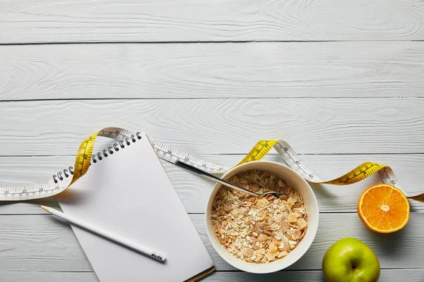 Top view of notebook with pencil, breakfast cereal in bowl, apple and orange near measuring tape on wooden white background with copy space — Stock Photo