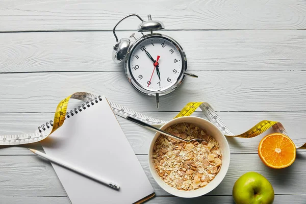 Vista dall'alto della sveglia, notebook con matita, cereali per la colazione in ciotola, mela e arancia vicino al metro su sfondo bianco in legno — Foto stock