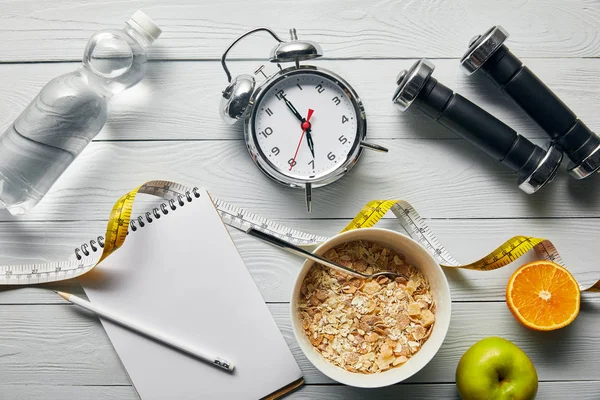Vista superior de despertador, mancuernas, agua, cuaderno con lápiz, cereales para el desayuno en tazón, manzana y naranja cerca de cinta métrica sobre fondo blanco de madera - foto de stock
