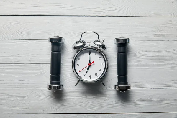 Flat lay with silver alarm clock and black dumbbells on wooden white background — Stock Photo