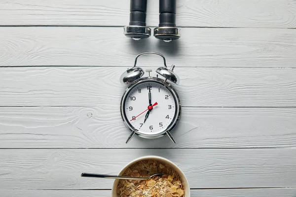 Tendido plano con despertador de plata, cereal de desayuno en tazón y pesas negras sobre fondo blanco de madera - foto de stock