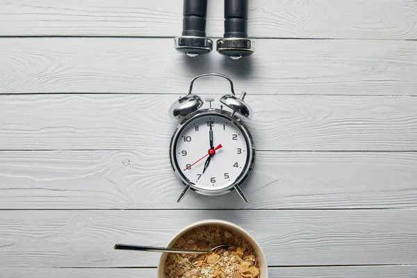 Flache Liege mit silbernem Wecker, Frühstücksflocken in Schüssel und schwarzen Hanteln auf weißem Holzgrund — Stockfoto