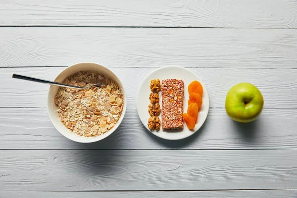 Plat étendu avec céréales pour petit déjeuner dans un bol, assiette avec des noix et des abricots secs près de pomme verte sur fond blanc en bois — Photo de stock