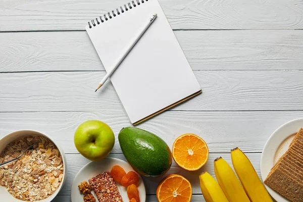 Верхний вид завтрака хлопья в миске, орехи, хрустящий хлеб и спелые фрукты возле ноутбука и карандаш на деревянном белом фоне — стоковое фото