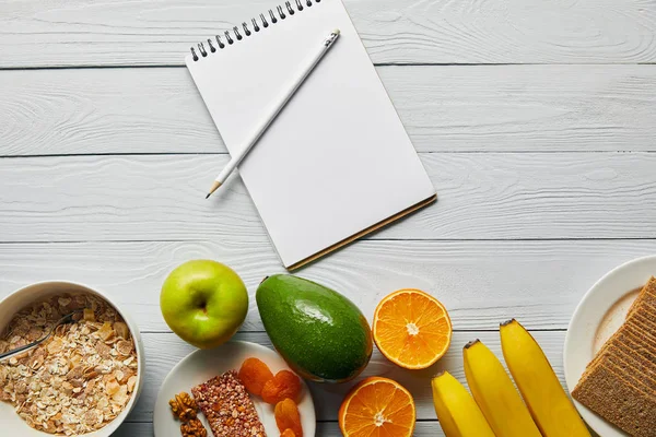 Top view of breakfast cereal in bowl, nuts, crispbread and ripe fruits near notebook and pencil on wooden white background — Stock Photo