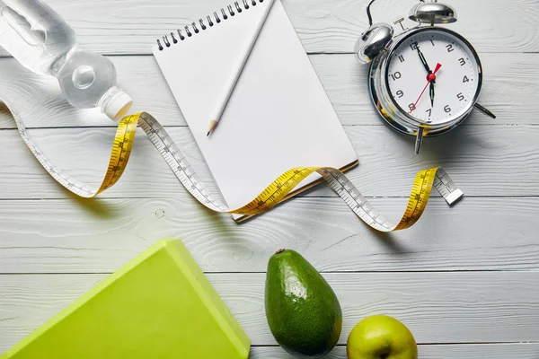 Top view of notebook and pencil, alarm clock, measuring tape, avocado, apple and water on wooden white background — Stock Photo