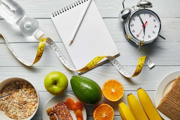 Top view of diet food near alarm clock, measuring tape, notebook and water on wooden white background — Stock Photo