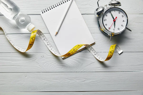 Vista dall'alto di nastro di misurazione, notebook in bianco, acqua e sveglia su sfondo bianco in legno con spazio di copia — Foto stock