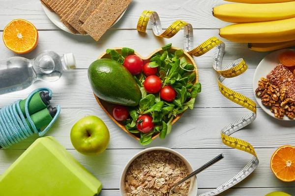 Top view of measuring tape, cereal, fruits and vegetables in heart-shaped bowl and sport equipment on wooden white background — Stock Photo