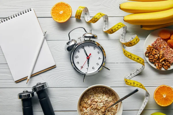 Ansicht von Maßband, Diätfutter und Kurzhanteln in der Nähe von blankem Notizbuch und Wecker auf weißem Holzgrund — Stockfoto