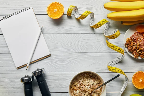 Vista superior de cinta métrica, pesas, alimentos dietéticos y cuaderno en blanco sobre fondo blanco de madera - foto de stock