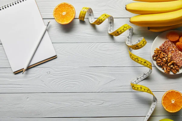 Vue du dessus du ruban à mesurer, des aliments diététiques et du carnet vierge sur fond blanc en bois avec espace de copie — Photo de stock