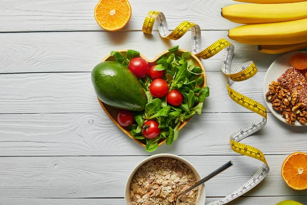 Draufsicht auf frisches Obst, Gemüse in herzförmiger Schüssel, Maßband und Müsli auf weißem Holzgrund — Stockfoto
