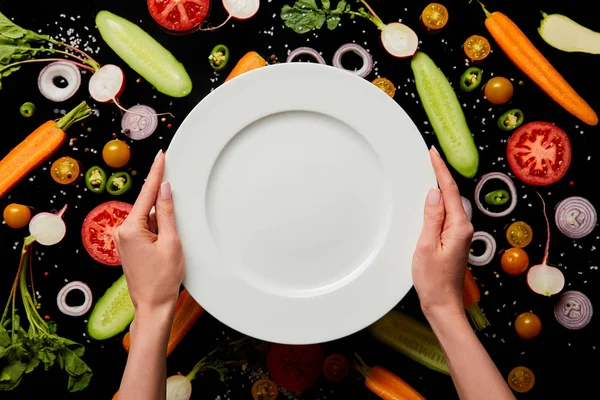 Cropped view of woman holding empty round plate on vegetable pattern background isolated on black — Stock Photo