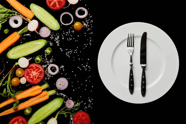 Top view of round plate with cutlery near vegetable slices isolated on black — Stock Photo
