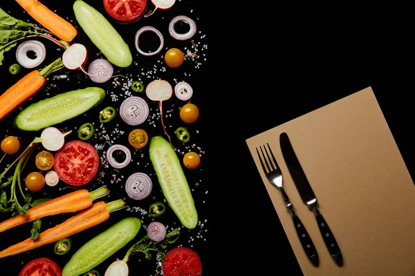 Top view of fresh vegetable slices with salt near paper blank and cutlery isolated on black with copy space — Stock Photo