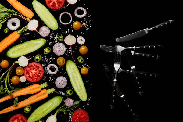 Top view of fresh vegetable slices with salt near knives and forks isolated on black with copy space — Stock Photo