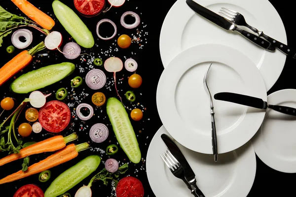 Vista superior de placas redondas y cubiertos cerca de verduras en rodajas aisladas en negro - foto de stock