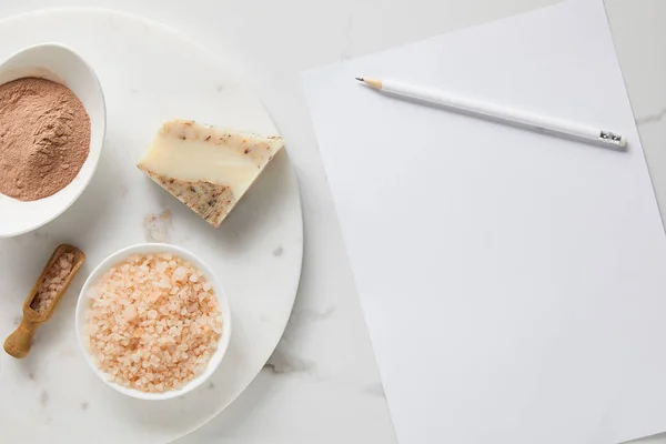 Верхний вид глиняного порошка, мыла и морской соли на мраморном столе рядом с белой бумагой и карандашом — стоковое фото