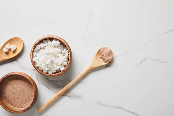 Top view of clay powder and sea salt in bowls on marble table near wooden spoons — Stock Photo