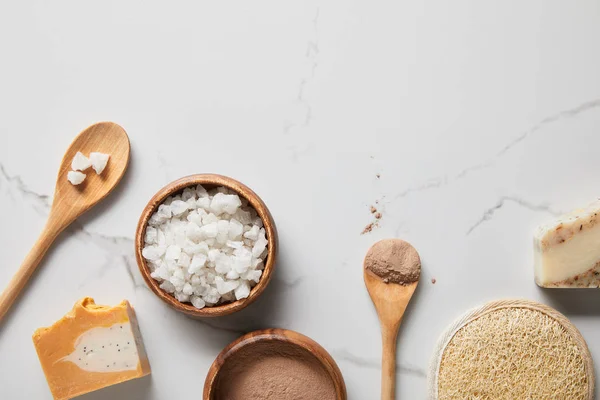 Top view of clay powder and sea salt in wooden bowls on marble table near spoons, soap and organic loofah — Stock Photo