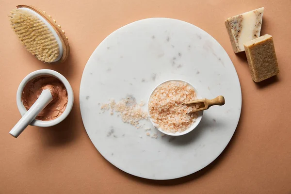 Top view of clay, wooden massage brush and soap on beige background near sea salt in bowl on marble circle — Stock Photo