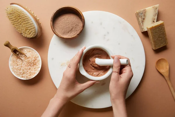 Vista recortada de la mujer mezclando arcilla en un tazón en círculo de mármol cerca de ingredientes cosméticos orgánicos y cepillo de masaje sobre fondo beige - foto de stock