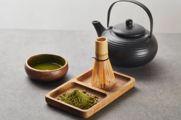 Selective focus of green matcha powder and bamboo whisk on wooden board near black teapot and bowl with tea — Stock Photo