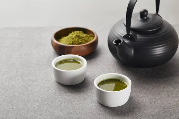 Selective focus of green matcha powder in wooden bowl near black teapot and white cups with tea on grey stone table — Stock Photo