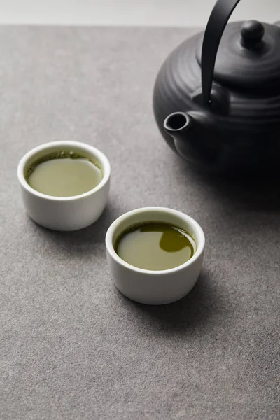 Selective focus of black teapot and white cups with matcha tea on grey stone table — Stock Photo