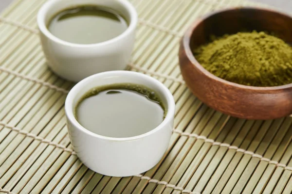 Foco seletivo de copos brancos com chá perto de pó matcha verde em tigela de madeira — Fotografia de Stock