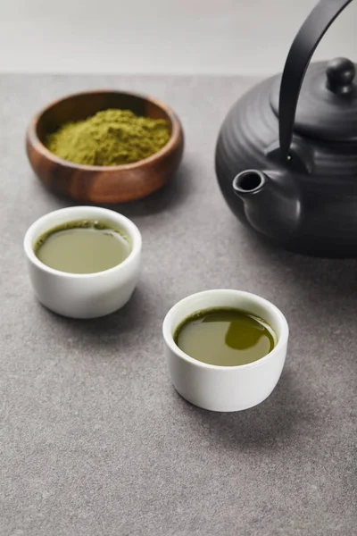 Selektiver Fokus von grünem Matcha-Pulver in Holzschüssel in der Nähe von Tassen mit Tee und schwarzer Teekanne — Stockfoto