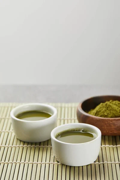 Fuoco selettivo di polvere di matcha verde in ciotola di legno vicino a tazze bianche con tè sul tappetino di bambù — Foto stock