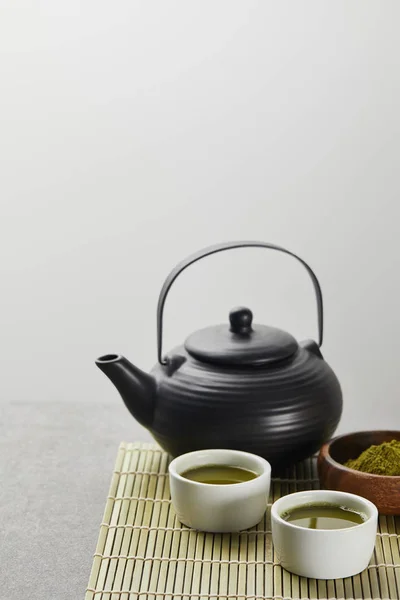 Selective focus of green matcha powder in wooden bowl near black teapot and cups with tea on bamboo table mat — Stock Photo