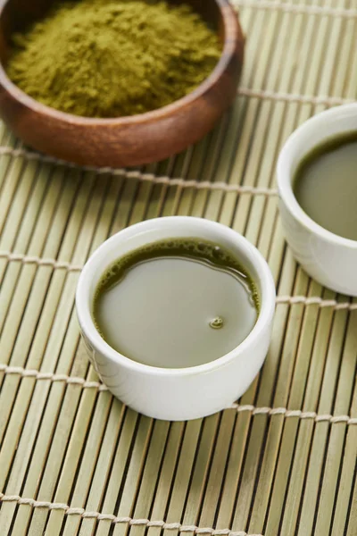 Selective focus of green matcha powder in wooden bowl near white cups with tea on bamboo table mat — Stock Photo