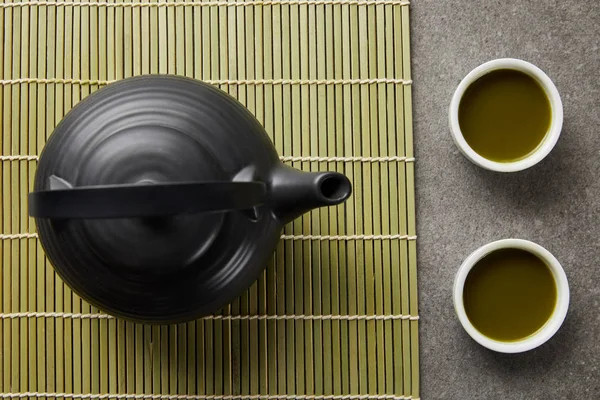 Vista superior de copos brancos com chá matcha verde perto de bule preto no tapete de mesa de bambu — Fotografia de Stock