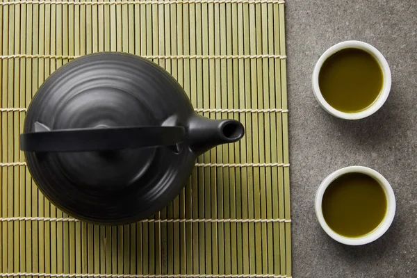 Vista dall'alto di tazze bianche con tè verde matcha vicino alla teiera nera sul tappetino di bambù — Foto stock