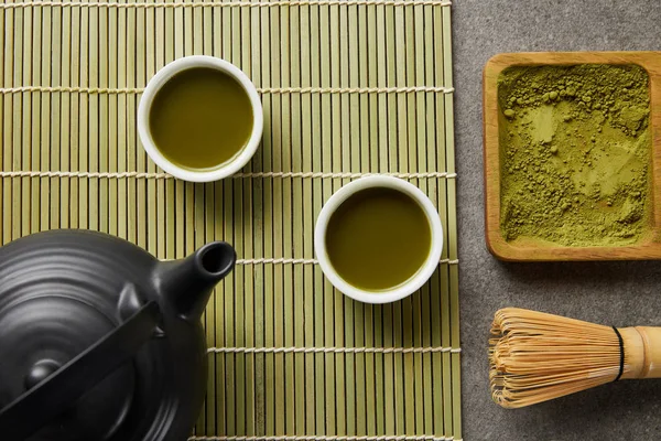 Vista dall'alto di tazze bianche con tè verde vicino alla teiera nera su tappetino in bambù con polvere di matcha su tavola di legno — Foto stock