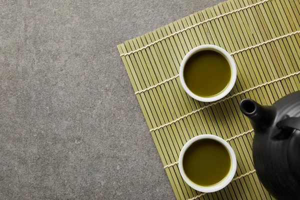 Вид сверху на белые чашки с зеленым чаем маття возле черного чайника на бамбуковом коврике — стоковое фото