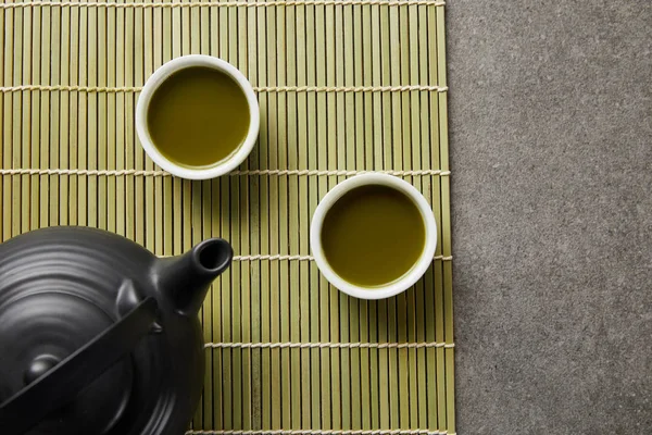 Vista dall'alto di tazze bianche con tè verde matcha vicino alla teiera nera sul tappetino di bambù — Foto stock