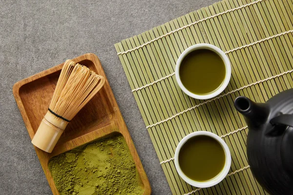 Vista superior de pó matcha verde com batedor de bambu em tábua de madeira perto de bule e xícaras brancas com chá verde no tapete de mesa — Fotografia de Stock