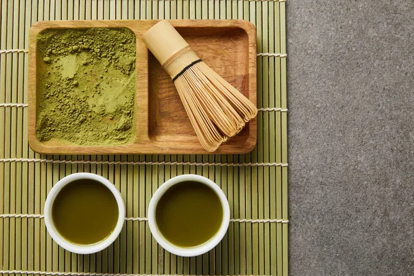 Ansicht von grünem Matcha-Pulver mit Bambusbesen auf Holzbrett in der Nähe weißer Tassen mit grünem Tee auf Tischmatte — Stockfoto