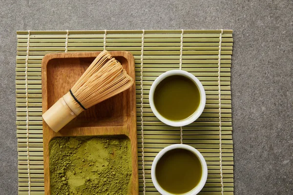 Vista superior de pó matcha verde com batedor de bambu em tábua de madeira perto de copos brancos com chá verde no tapete de mesa — Fotografia de Stock