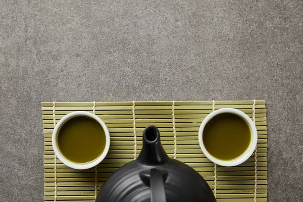 Vista dall'alto di tazze bianche con matcha e teiera nera su tappetino di bambù verde — Foto stock