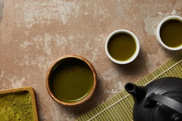 Vue du dessus de la poudre de matcha, bol et tasses avec du thé vert sur la surface vieillie près du tapis de table en bambou et théière noire — Photo de stock